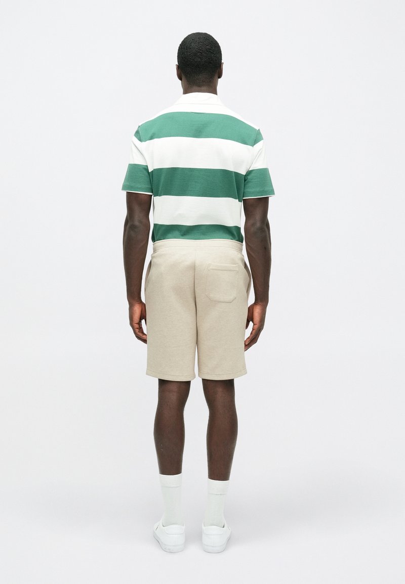 Groen en wit gestreepte polo met beige shorts. De shorts hebben een achterzak. Witte instap schoenen en lichte sokken worden gedragen.