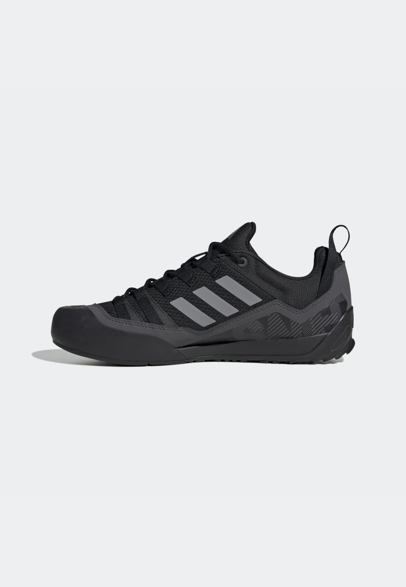Chaussure de sport Adidas noire et gris foncé avec un design bas, trois bandes grises sur le côté, fermeture à lacets et languette au talon.