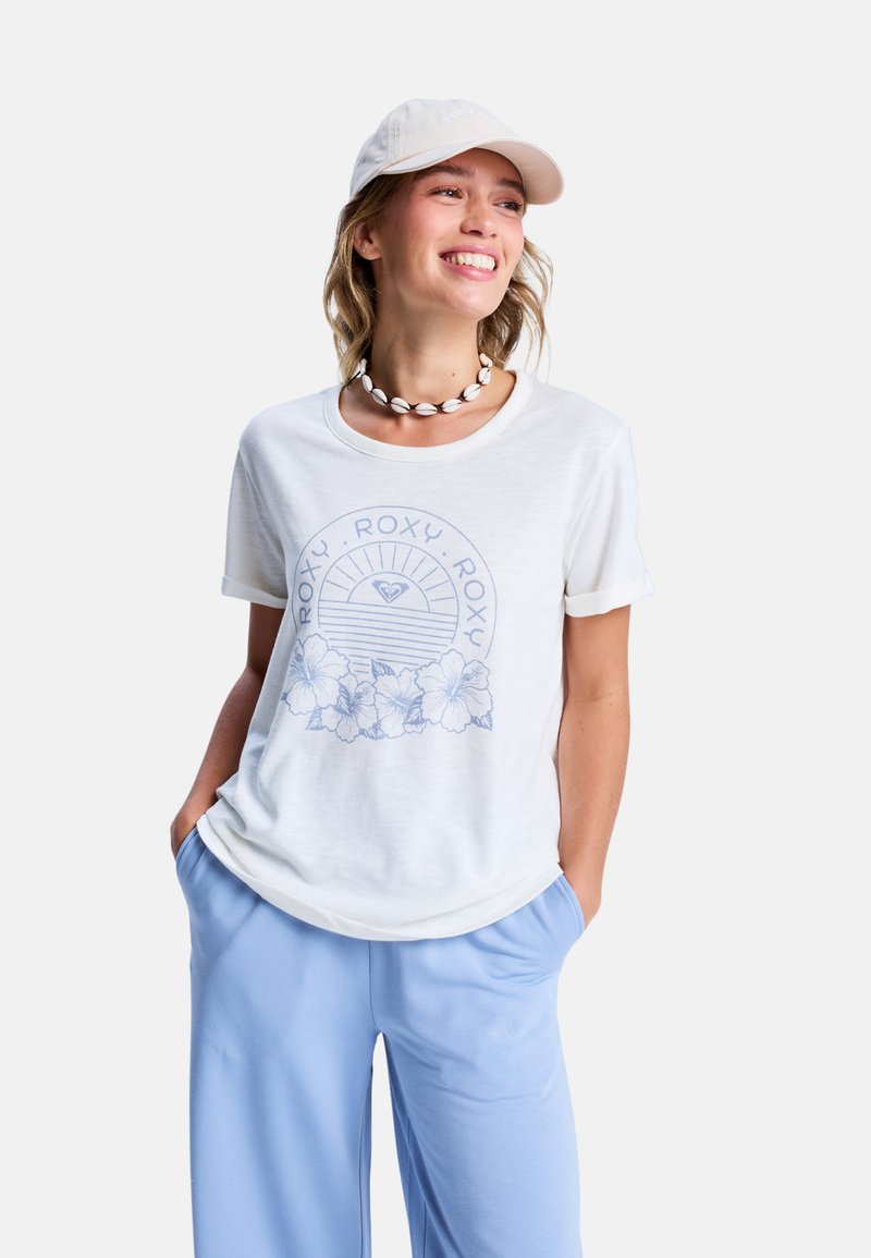 Witte t-shirt met blauw bloemenmotief en vintage tekst, korte mouwen en een relaxte pasvorm, gecombineerd met lichtblauwe broeken.