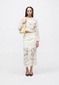 Witte gebloemde kanten cropped top met geschulpte randen, gecombineerd met een bijpassende nauwsluitende rok. Model houdt een gele tas vast en draagt bordeauxrode hakken.