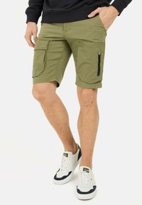 Olivgrüne Cargo-Shorts mit mehreren Taschen, einem lockeren Schnitt und einem strukturierten Stoff. Kombiniert mit weißen und marineblauen Sneakers.