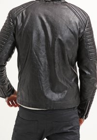 Giacca di pelle nera con pannelli imbottiti sulle spalle e design aderente. Presenta cerniere sulle maniche e una superficie liscia e leggermente testurizzata.