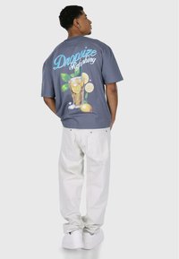 Grå bomulds t-shirt med et farverigt drikkevaremotiv på ryggen. Parret med hvide bukser og hvide sneakers. Løs pasform og korte ærmer.