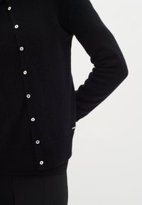 Musta neulecardigan pyöreällä kaula-aukolla, nappikiinnitys edessä ja hopeanväriset napit. Sisältää ribbigolfin helman ja pienen metallisen yksityiskohdan sivussa.