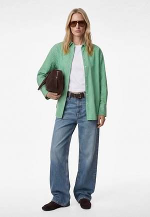 Kobieta w zielonej koszuli w paski z guzikami, białym T-shircie, szerokich dżinsach, brązowych butach, w okularach przeciwsłonecznych, trzymająca pod pachą brązową torebkę.