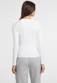 Camicia bianca a maniche lunghe con scollo rotondo, tessuto morbido e design aderente, abbinata a pantaloni della tuta grigi chiari.