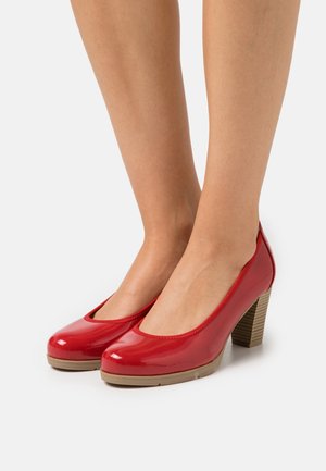 Jambes portant des chaussures à talons hauts rouges brillants, à bouts arrondis et talons en bois empilés, sur un fond blanc.