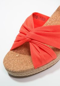 Gros plan sur une sandale avec une semelle en liège et une large bordure en jute tressée, avec une bride torsadée en tissu corail vif sur la semelle.