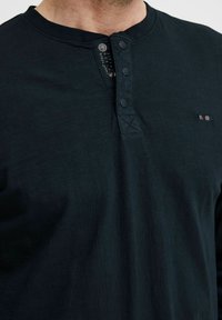 Tume roheline pikkade varrukatega henley särk, mis on valmistatud tekstuurilisest kangast. Omab kolme nupuga rinnaosa ja väikest triibulist aktsenti rinnal.