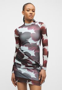 Γυναίκα που φοράει μακρυμάνικο, εφαρμοστό φόρεμα με μεγάλο μπορντό και γκρι ανθικό σχέδιο και έναν δεμένο κόμπο στη αριστερή πλευρά της μέσης.