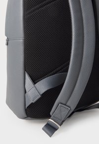 Gros plan d'un sac à dos gris avec un panneau arrière en maille noire rembourrée et une sangle d'épaule grise réglable avec boucle métallique.