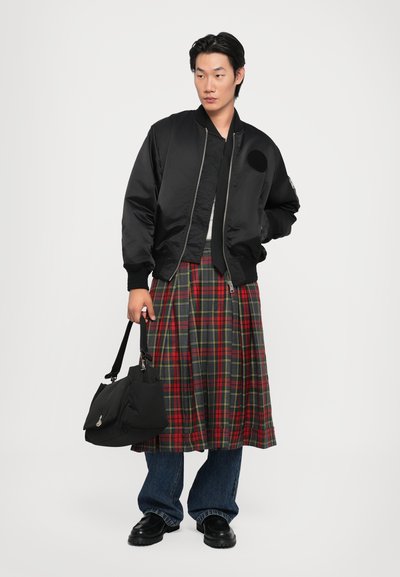 Schwarze Bomberjacke über einem weißen Hemd, kombiniert mit einem rot-grünen Karorock. Dunkle Jeans und schwarze Schuhe. Hält eine schwarze Tasche.