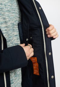 Navy outerwear jacket with a zip front, snap closures, and an interior pocket, featuring an orange care label and textured gray sweater underneath.