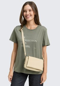 Donna con lunghi capelli castani che indossa una maglietta grafica verde oliva, jeans scuri e una borsa a tracolla beige, che sorride leggermente di lato.
