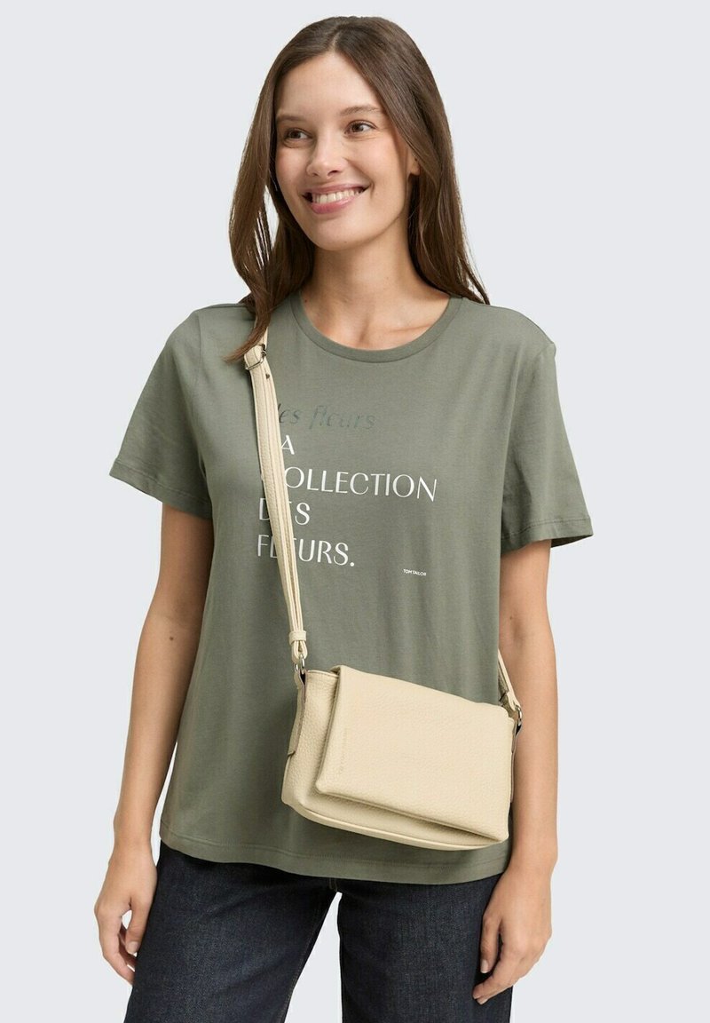Donna con lunghi capelli castani che indossa una maglietta grafica verde oliva, jeans scuri e una borsa a tracolla beige, che sorride leggermente di lato.