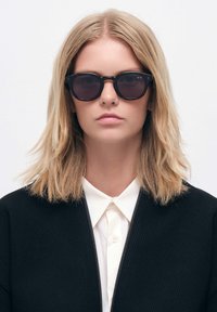 Aurinkolasit, joissa on tummat linssit, pyöreät kehykset ja kiiltävä musta pinta. Mallilla on päällään musta teksturoitu takki valkoisen paidan päällä.