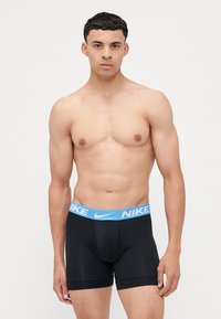 Čierne boxerky s modrým pásom s logom Nike, so priliehavým strihom a hladkou textúrou, vystavené na mužskom modeli.