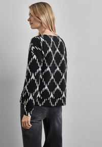 Sort tekstureret sweater med et hvidt diamantmønster, med rund halsudskæring og lange ærmer, båret med mørkegrå bukser.