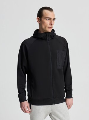 Uomo con capelli pettinati all'indietro che indossa una felpa nera con cerniera e tasca sul petto, in piedi davanti a uno sfondo grigio chiaro uniforme, guardando verso destra.