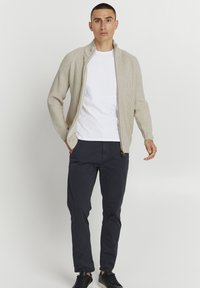 Cárdigan de punto beige con cremallera y cuello alto, con textura de canalé, llevado sobre una camiseta blanca, combinado con pantalones azul marino oscuros y zapatillas negras.