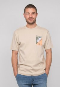 T-shirt en coton beige à manches courtes, col rond, avec un design de palmier imprimé sur la zone de la poche avant. Coupe décontractée, texture lisse.