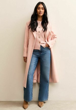 Manteau rose clair ample porté sur un pull rose tricoté à câbles avec des boutons en forme de fleur, associé à un jean bleu à jambes larges et des talons pointus beiges.