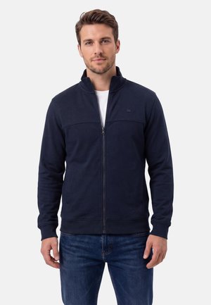 Uomo con giacca blu navy con zip e jeans blu, in piedi contro uno sfondo chiaro e uniforme, rivolto verso l'obiettivo con un'espressione neutra.