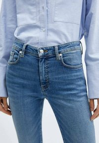 Jeans de mezclilla azul claro con un diseño ajustado, cinco bolsillos y un cierre de botón en la cintura. Un ligero desgaste añade textura.