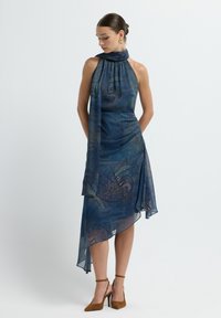 Vestido con cuello halter en un patrón de paisley azul y púrpura, con un dobladillo asimétrico y tejido translúcido. Combinado con zapatos de tacón alto marrones.