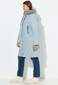 Lunga giacca azzurro chiaro con cappuccio in peluche marrone. Presenta tasche con zip nere e una vestibilità comoda. Abbinata a jeans scuri e stivaletti crema.