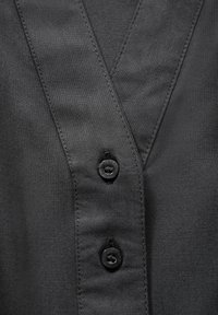 Camicia nera con colletto a V, tessuto liscio e due bottoni neri sulla parte anteriore. Cuciture dettagliate visibili.