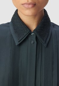 Blusa nera con colletto strutturato e decorazioni di perline, pieghe verticali e chiusura frontale con bottoni. Finitura in tessuto liscia.