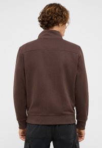 Brown sweatshirt with a high collar, ribbed cuffs, and hem. Smooth fabric with a simple design and a subtle seam detail across the shoulders.