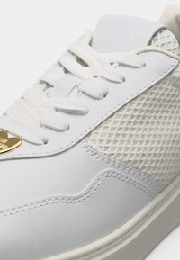 Λευκό αθλητικό παπούτσι με πλαϊνές πλευρές από υφαντό δίχτυ, λευκά κορδόνια, λεπτομερείς ραφές και ένα μικρό χρυσό μενταγιόν σε σχήμα καρδιάς κοντά στη μύτη.