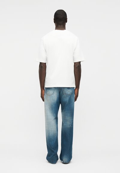 Άνδρας που στέκεται με την πλάτη προς τη θέα, φορώντας ένα απλό λευκό κοντομάνικο μπλουζάκι και ξεβαμμένα μπλε τζιν με ελαφρώς φθαρμένα τελειώματα.