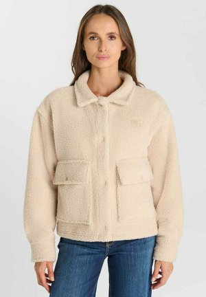 Frau trägt eine beige flauschige Jacke mit großen Fronttaschen und blaue Jeans, steht vor einem schlichten hellgrauen Hintergrund.