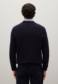 Maglione blu navy a maglia con finitura texture, polsini e orlo a costine e scollatura rotonda, visto da dietro. Camicia azzurra con colletto visibile.
