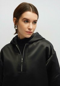 Felpa nera con cappuccio e zip parziale, realizzata in tessuto liscio. Presenta un colletto alto e maniche larghe. Dettaglio della zip in argento.