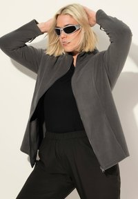 Veste en polaire grise avec fermeture éclair à l'avant, col montant et poches latérales, portée sur un haut noir et associée à un pantalon noir.