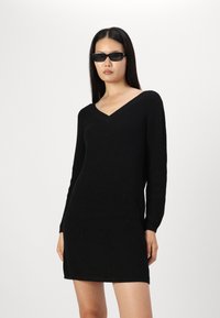 Abito nero lavorato a maglia con scollo a V e maniche lunghe, caratterizzato da una trama a coste e una silhouette aderente, fino al ginocchio. Accompagnato da occhiali da sole scuri.