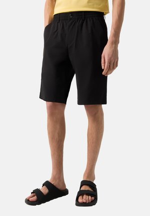 Mann trägt schwarze Shorts mit elastischem Bund, schwarze Sandalen mit Riemen und ein gelbes Hemd, steht mit entspannten Händen seitlich am Körper.