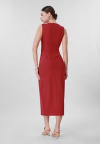 Γυναίκα με πλάτη, φοράει κόκκινο midi φόρεμα χωρίς μανίκια, εφαρμοστό, με μπεζ σανδάλια με τακούνι και χρυσά σκουλαρίκια και βραχιόλι σε απλό φόντο.