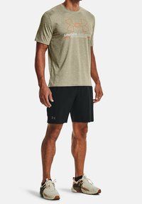 Maglietta sportiva Under Armour in oliva con logo arancione, abbinata a pantaloni corti neri e scarpe sportive chiare, con dettagli a fantasia.