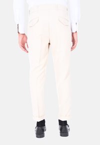 Pantaloni beige su misura con vestibilità slim, dotati di due tasche posteriori con chiusure a bottone e risvolti, abbinati a scarpe eleganti nere.