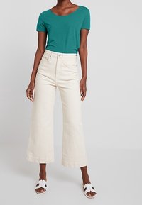 T-shirt teal a maniche corte, jeans a gamba larga bianchi a vita alta e sandali bianchi. Tessuto di cotone morbido, vestibilità comoda e orli dritti.