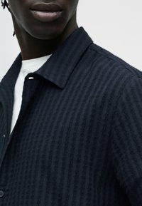 Gros plan d'une personne portant une chemise à boutons texturée bleu marine par-dessus un sous-vêtement blanc, montrant les lèvres et le bas du visage, sans détails en arrière-plan.