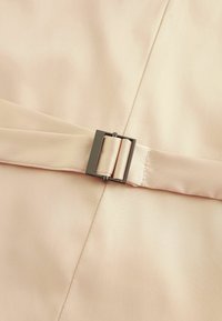Un tissu beige clair présente une boucle en métal argenté élégante, cintrant une sangle lisse. La texture est douce, avec un léger éclat.