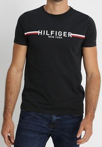 Černé bavlněné tričko s bílým a červeným horizontálním pruhem a textem "HILFIGER NEW YORK". Krátké rukávy a design s kulatým výstřihem.