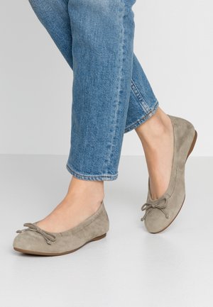 Fødder iført taupe ruskind ballet flats med sløjfedetalje kombineret med blå denimjeans mod en ensfarvet lys baggrund.