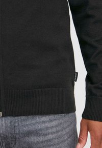 En närbild som visar den nedre delen av en person som bär en svart tröja och grå jeans, med en synlig klädesetikett på tröjan.